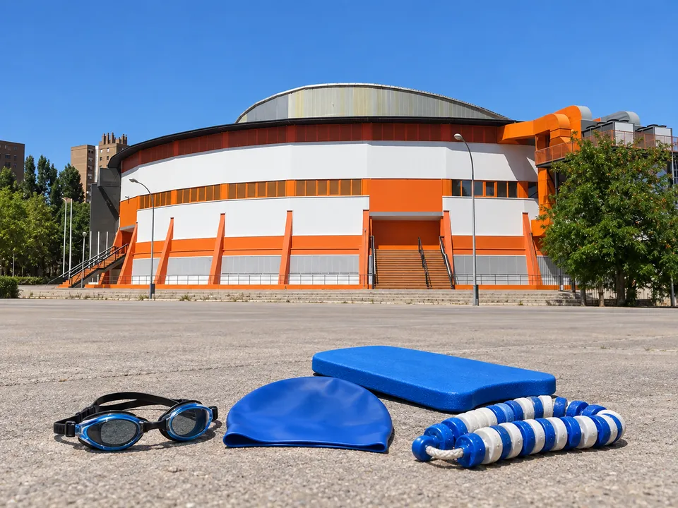 AI-generated illustration: empty Font de Sant Lluís arena exterior with swim goggles, cap, kickboard and a lane-rope segment.