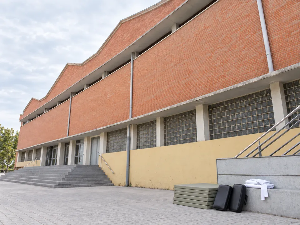 AI-generated illustration: empty Cabanyal-Canyamelar sports hall exterior with tatami mats, focus pads and a folded white karate uniform.