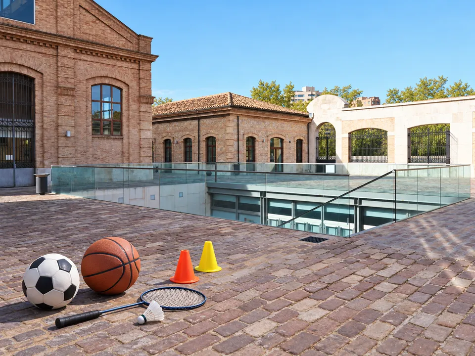AI-generated illustration: empty Petxina courtyard with football, basketball, cones, badminton racket and shuttlecock.